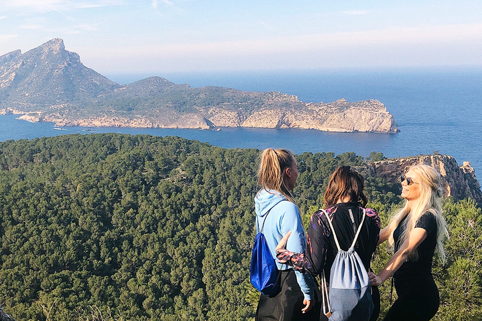 Tre unga kvinnor står på en höjd och blickar ut över ett vackert landskap av skog, berg och hav. Foto.