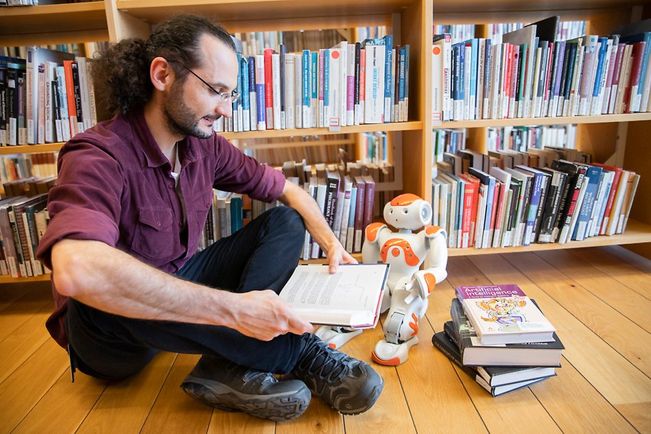 Man sitting on the floor of a library reading, a small humanoid robot is standing next to him. Photo.