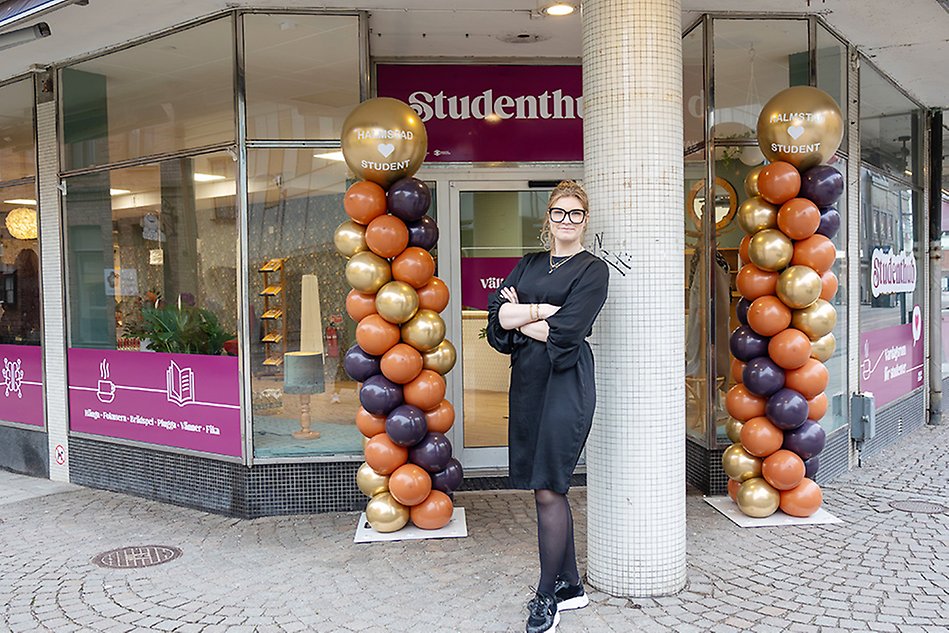 A young women with bright hair and glasses is standing infront of a building with an entrance and a door behind her.