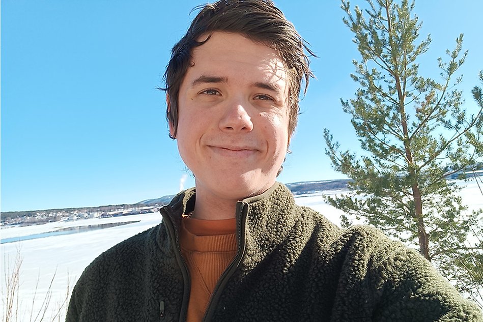 En man står utomhus i en grön fleecetröja och tar en selfie. I bakgrunden finns en frusen sjö, skog och klarblå himmel. Foto.