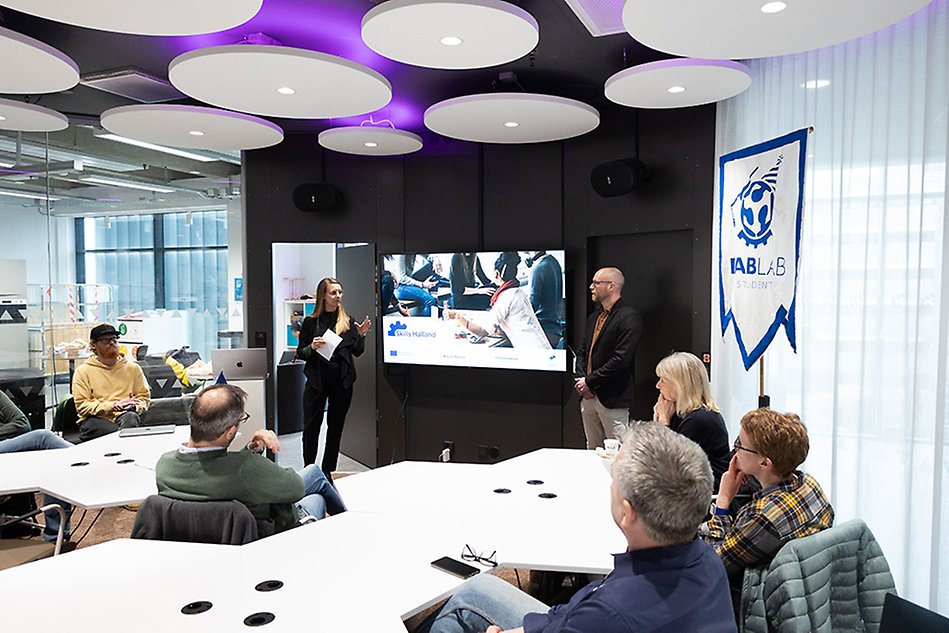 Kvinna och man står framför en PowerPoint-presentation. Deltagare sitter vid bord och lyssnar på dem. Foto.