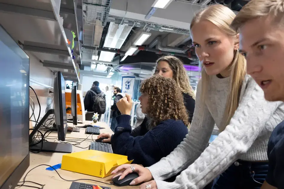 Fyra studenter sitter vid två datorer och ser fokuserade ut. I bakgrunden går studenter med ryggsäckar i motsatt riktning.