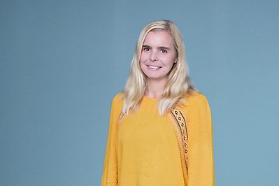 Blond kvinna står och ler mot kameran framför blå vägg. Foto.