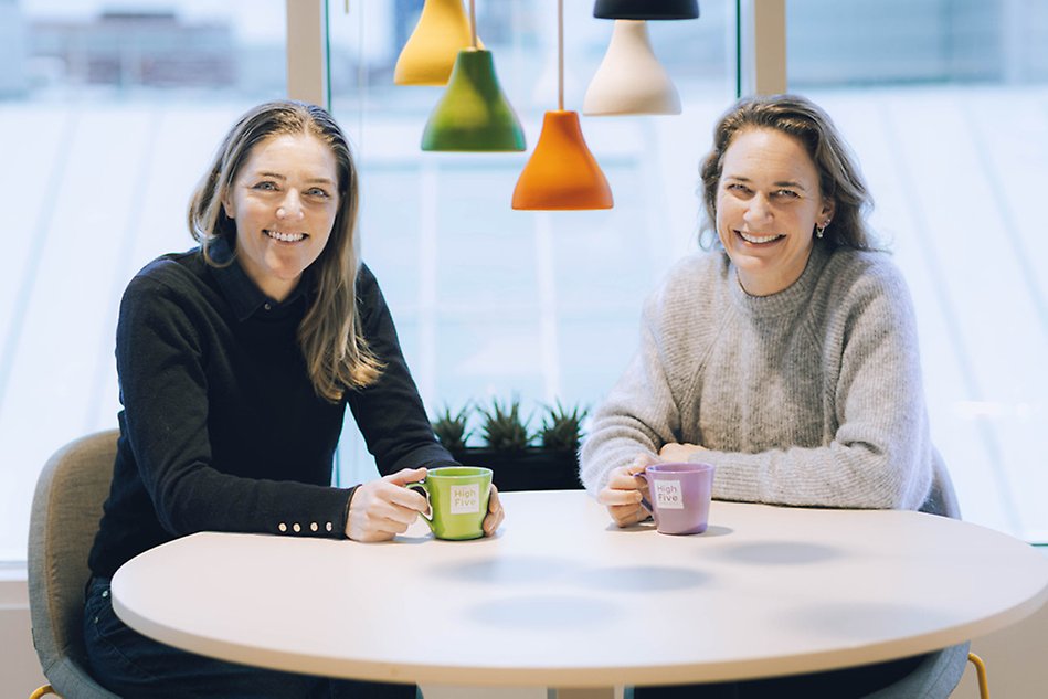 Två leende kvinnor sitter vid ett runt bord med kaffekoppar i sina händer. Kvinnan till vänster har en svart tröja och kvinnan till höger en grå tröja. Foto.