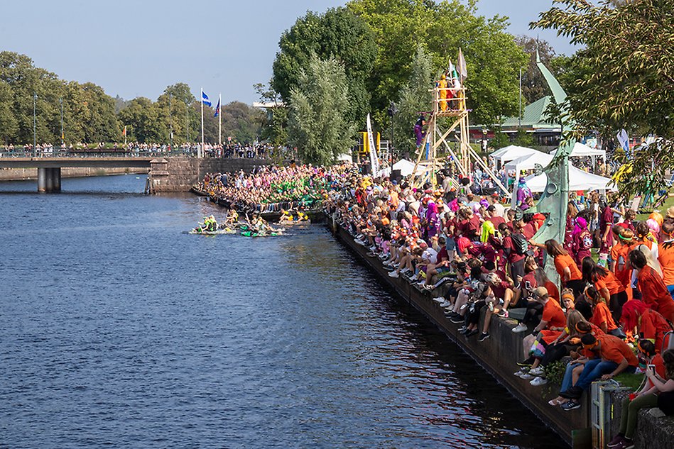 Bild över å med bro i bakgrunden och mycket folk i färgglada kläder på åns högra sida. Tre flottar i ån. Foto.