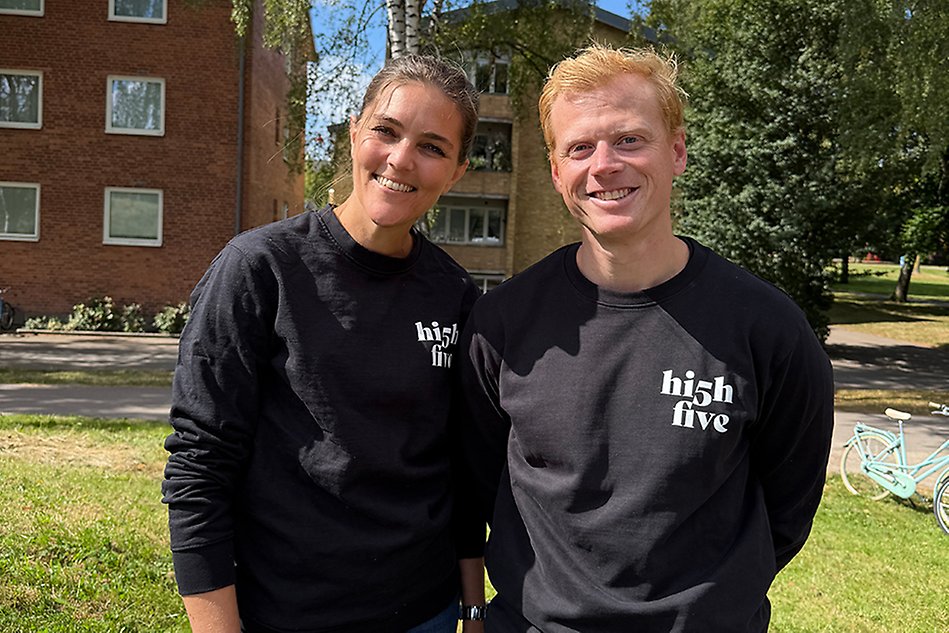 Två personer utomhus ler mot kameran. De bär matchande svarta tröjor. Foto.