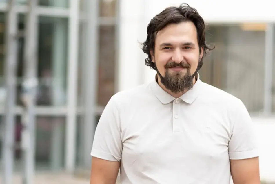 Student i brunt hår och skägg ler mot kameran. Han bär en ljusbeige pikétröja utomhus. Foto.