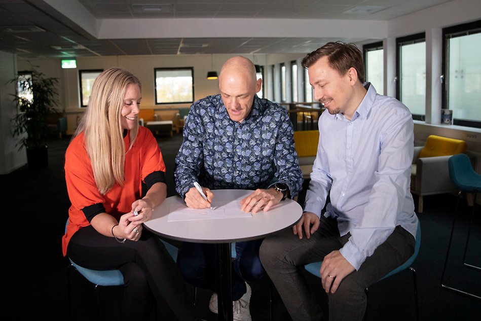 Three persons sit around a table. The person in the middle writes on a paper.