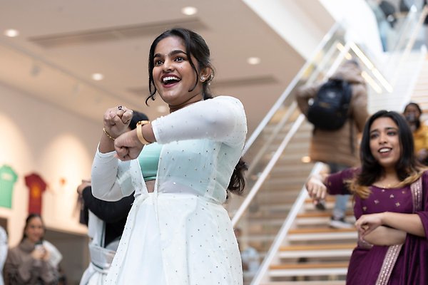 Person som ler stort och dansar. Hon har färgglada indiska kläder på sig och i bakgrunden syns fler som dansar. Foto.