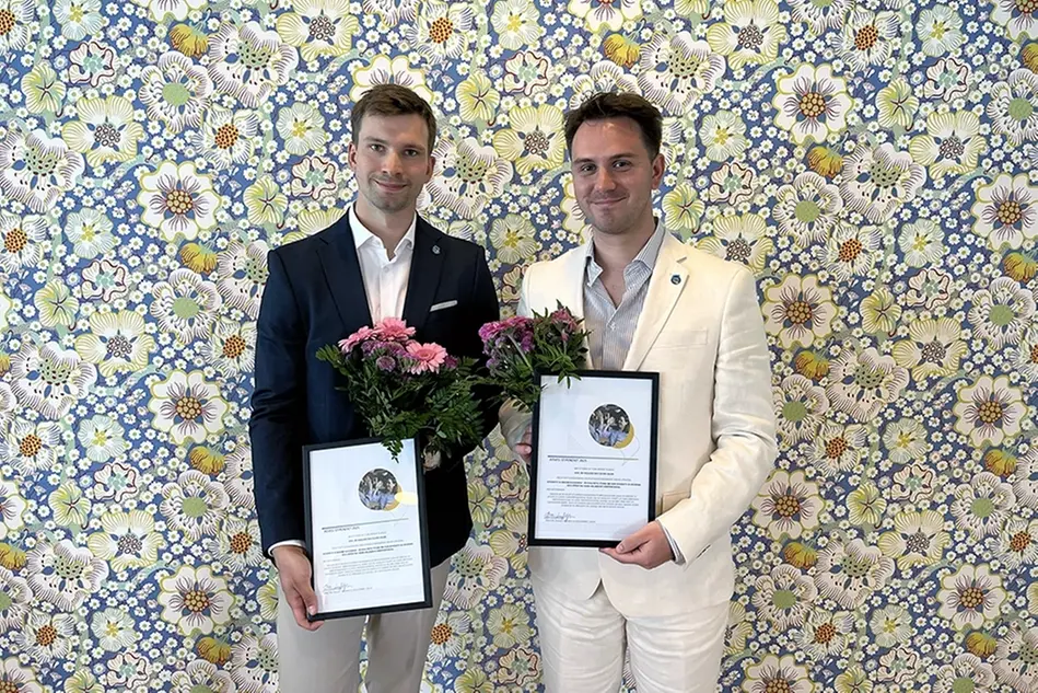 Två personer i kostym håller i diplom och blommor för att de fått Forterros stipendium. Foto.