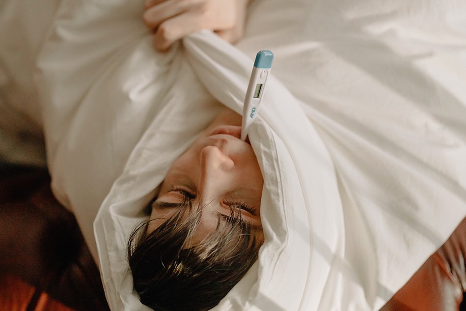 En person ligger ned, inbäddad i ett tjockt täcke och med en febertermometer i munnen. Foto.
