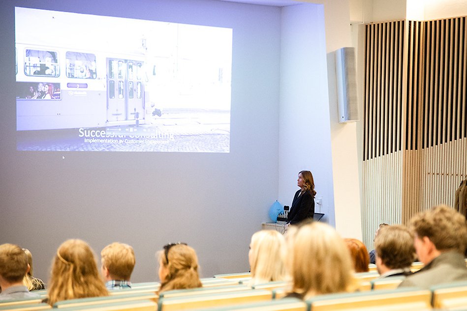 En föreläsningssal full med personer och en kvinna som presenterar. Foto.