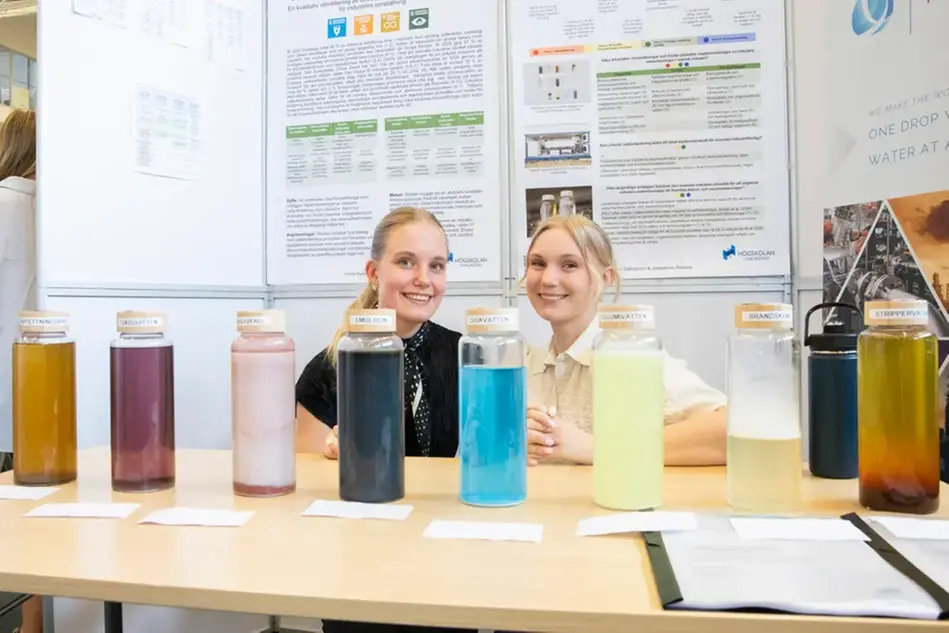 Två studenter ler och hukar bakom sitt bord. På bordet står cylindrar fyllda med vätskor i olika färger.