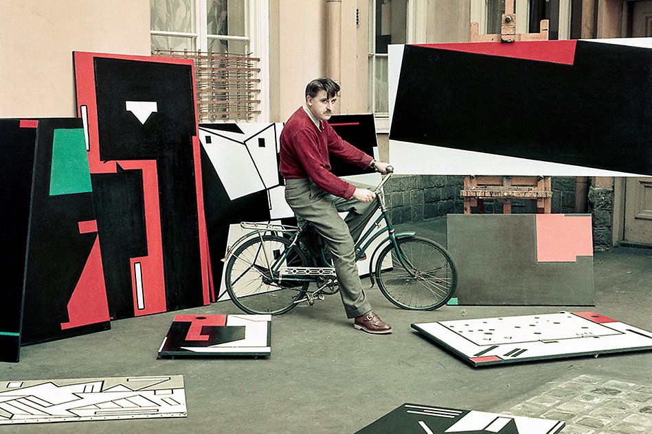 Konstnären Olle Baertling sitter på en cykel omgiven av sina abstrakta konst.
