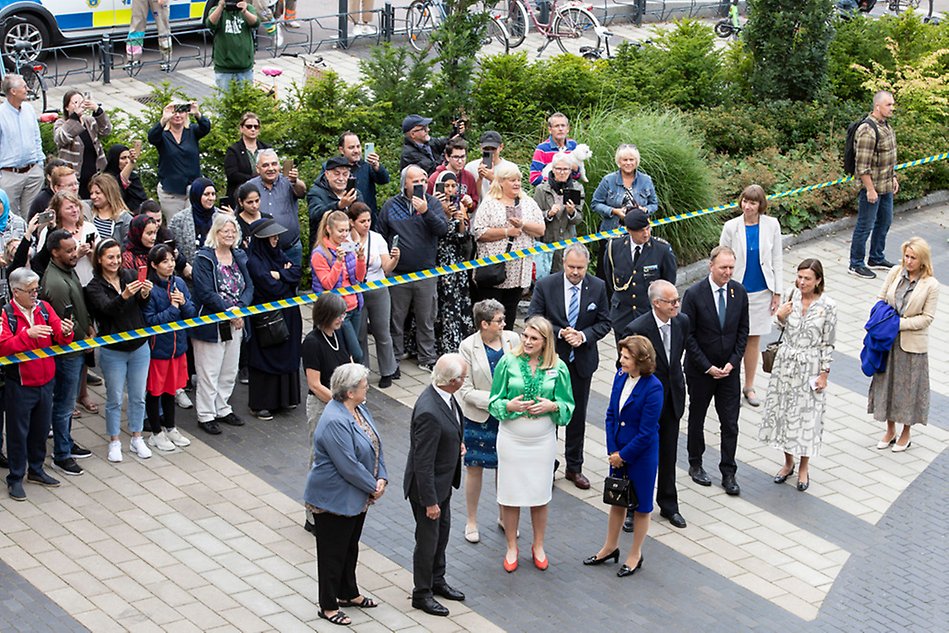 Seen from above in the background a large crowd behind cordon. In the foreground a number of people standing and talking in a group. Photo