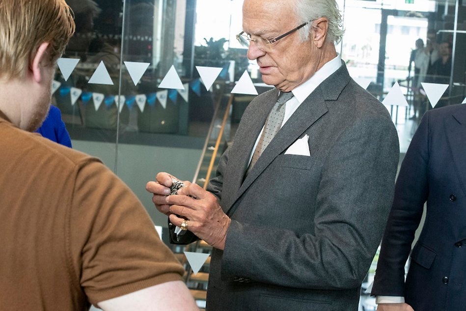A man in a suit (King of Sweden) stands to the right and looks at a small gadget that he holds in his hands. To the left is the back panel of a man.