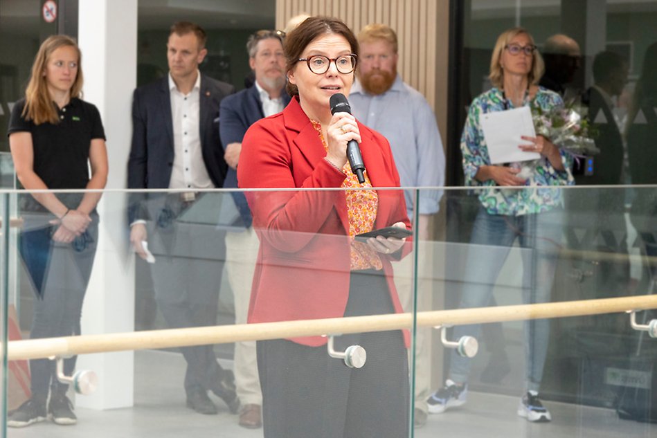 Kvinna i brunt uppsatt hår med röd kavaj och glasögon talar i en mikrofon. I bakgrunden står folk och lyssnar. Foto.