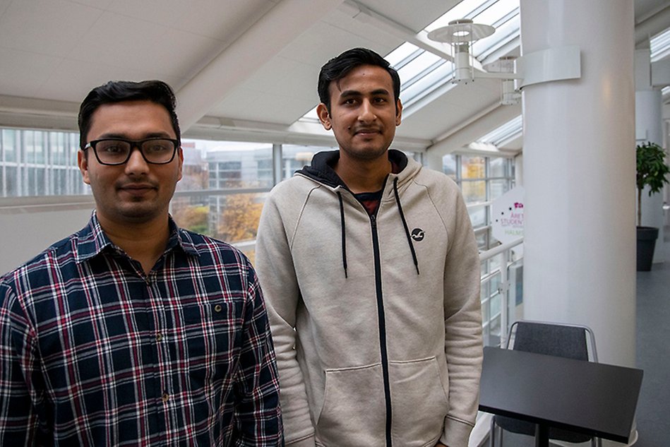 Two men with dark hair standing in a corridor with glass walls behind them. Photo.