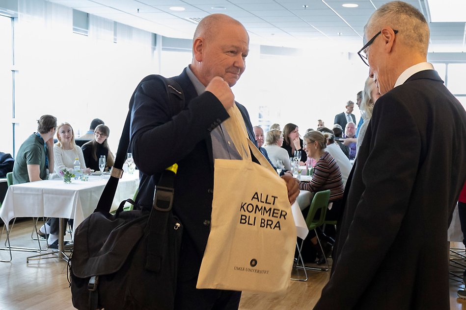 I förgrunden står två män står mittemot varandra och den ena håller upp en kasse som det står Allt kommer att bli bra på. I bakgrunden sitter flera personer vid bord. Foto