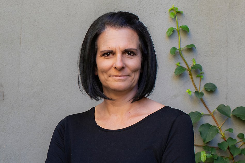 A dark haired woman looks into the camera and smiles. In the background a twig from a plant.