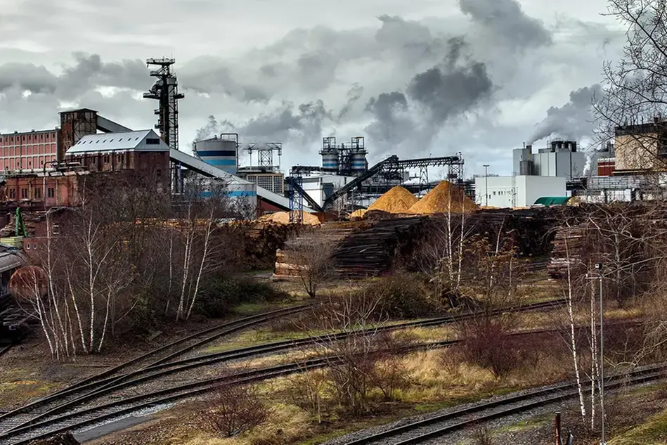 Industrial site with woodchip piles, factory buildings and smoke, next to railway tracks.