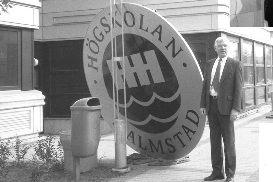 Man standing outside, next to a large sign with the old Halmstad University logo on it. Black and white photo.