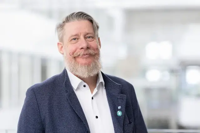 Man i vitt hår, skägg och mustasch ler mot kameran. Han bär en vit skjorta och denimblå kavaj. I bakgrunden finns en ljus korridor.