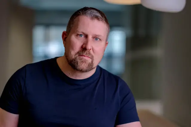 En kortklippt man i skägg sitter ner och lutar sig mot ett bord. Han bär en mörkblå t-shirt och tittar mot kameran. 