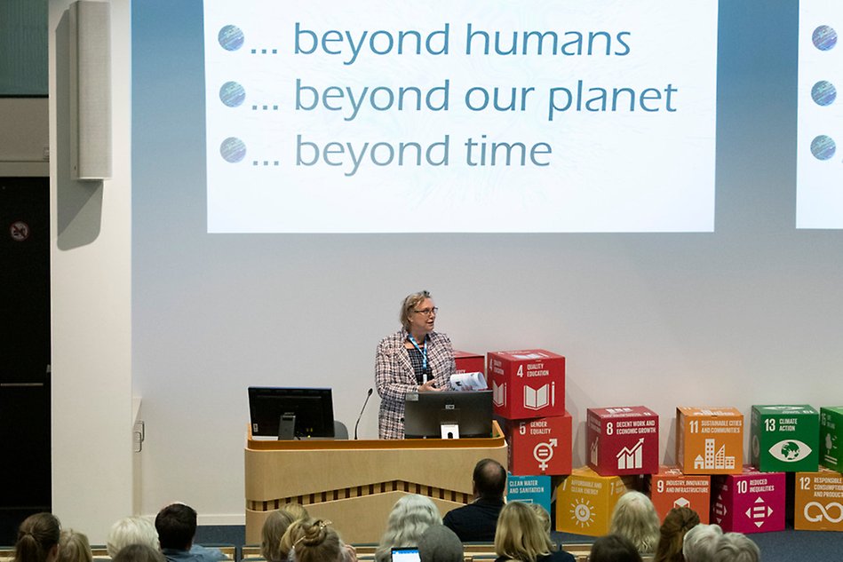 Woman in dress jacket is standing in front of a presentation where it says "Beyond humans, beyond the planet, beyond time". Photo.