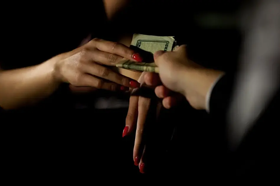 Två personer som utbyter en bunt dollarsedlar. Den ena handen har rött nagellack. Den andra ser ut att ha kostymklädd arm. Foto
