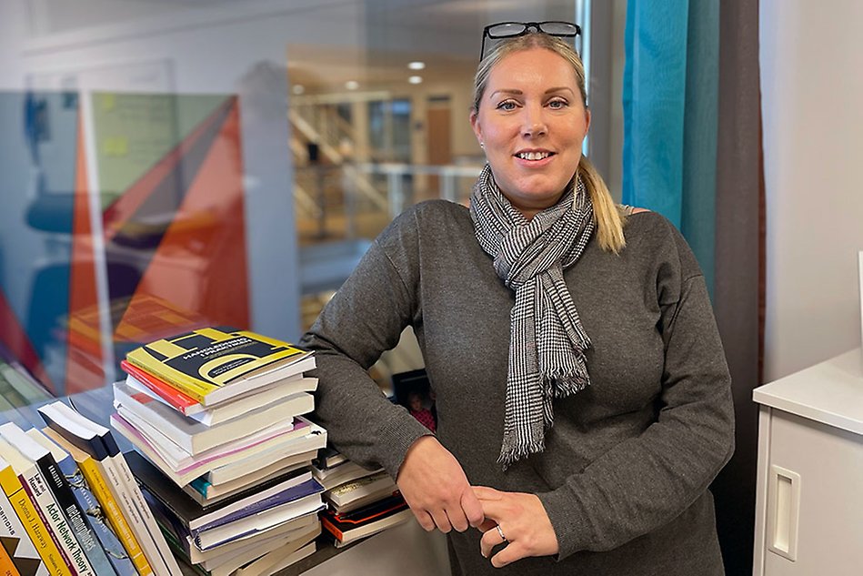Kvinna som ser glad ut står inomhus och lutar sig mot en hög med böcker