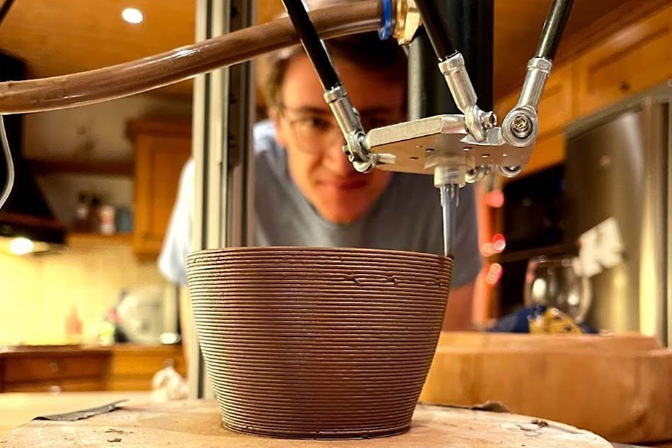 A 3D printer prints a pot. A person is standing and watching in the background. Photo.