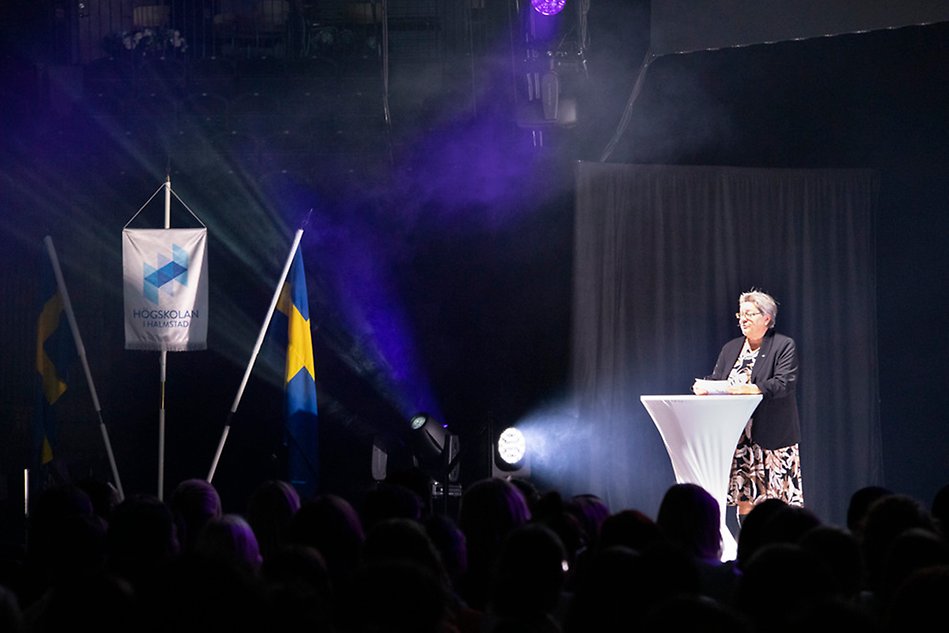 En kvinna står på en scen vid ett podium med strålkastarljus på sig. Till vänster om henne syns en vit flagga med Högskolans i Halmstads logotyp och två svenska flaggor. Foto.
