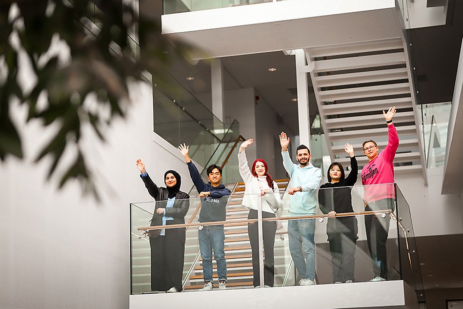 A group of students smiling and waving on a balcony at campus. Photo.