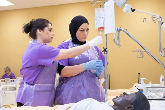 Två sjuksköterskor tittar på ett dropp och i sängen ligger en docka