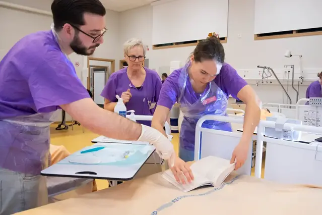 Två sjuksköterske studenter tittar i en bok och en lärare står bakom i sjukhus miljö
