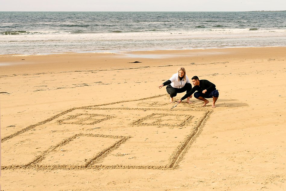 Bild på två unga studenter som sitter på huk och ritar hus i sanden. Fotografi