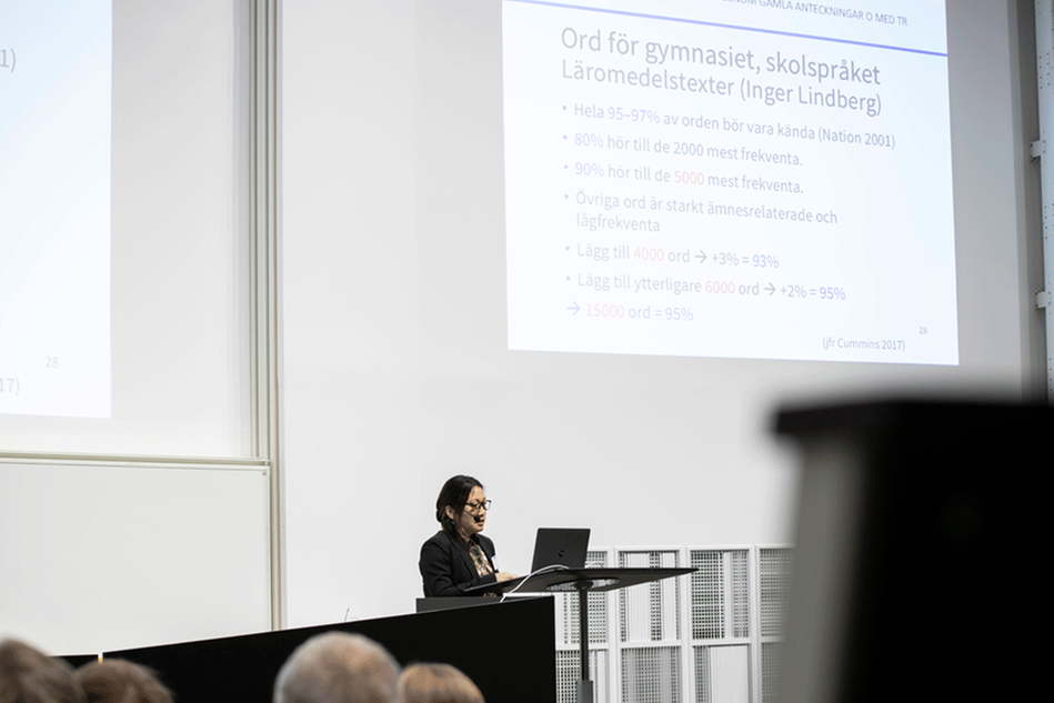 Kvinna i svart hår och svart kavaj, syns på podium med power point-bild bakom sig.