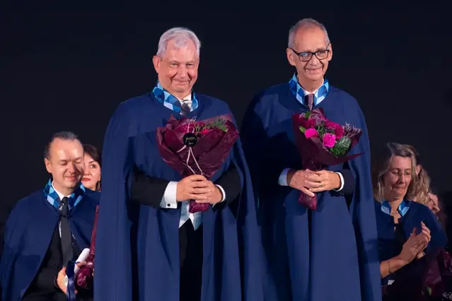 Två personer i caper med blommor i händerna står  framför andra personer som sitter. Foto 