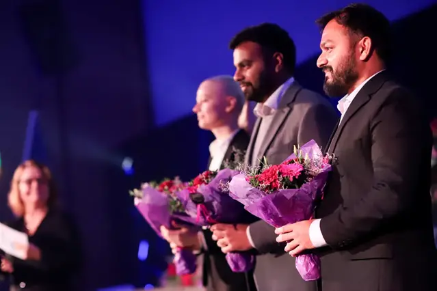 En grupp personer står och sitter på en scen och håller blommor i sina händer. Foto