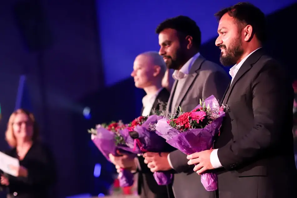 A group of people are standing and sitting on a stage, holding flowers in their hands. Photo