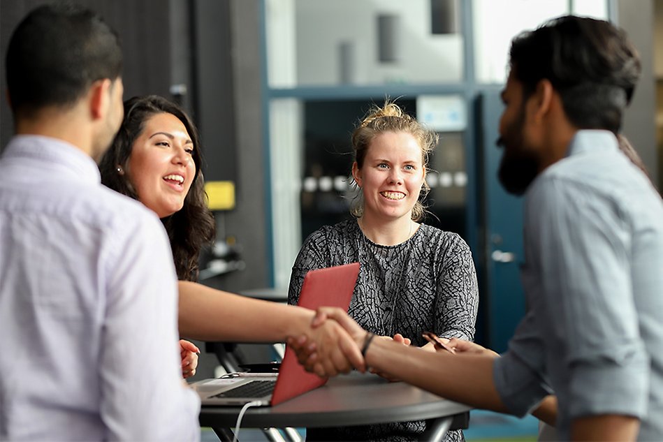 Four people standing around a table, two of them shaking hands across the table. Photo.