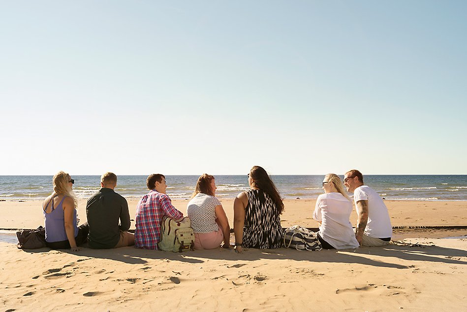A group of people sitting on a beach, looking towards the ocean. Photo.