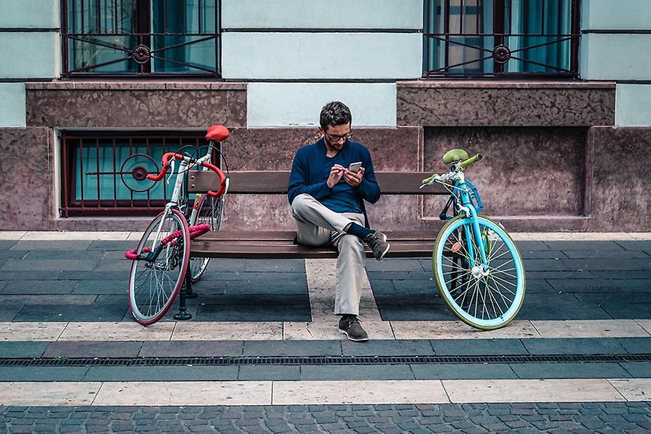 A man sitting on a bench in a street, a bike is leaning against each side of the bench. Photo.