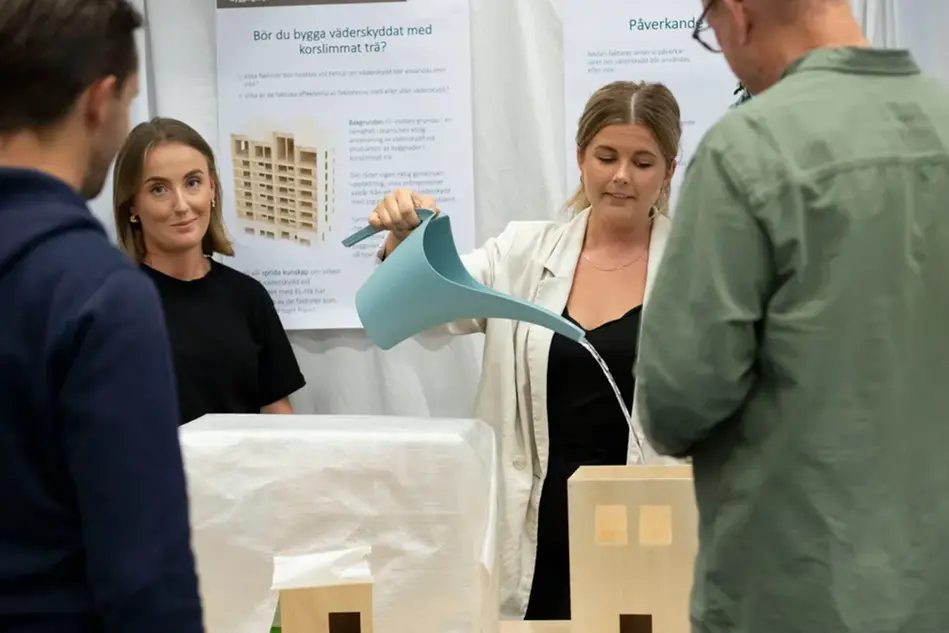 Två studenter står och pratar med två mässbesökare. Den ena studenten visar deras arbete. Foto.