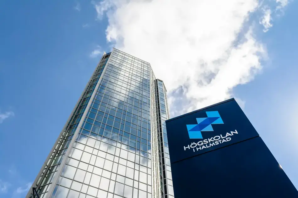 View of blue sky with a tall building and a column with the Halmstad University logo on it against the sky. Photo.