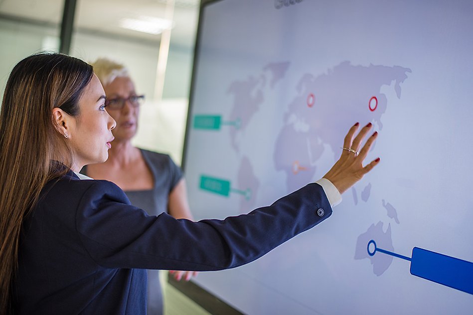 Two people looking at a world map on smart screen. One of them is explaining something to the other. Both are women. Photo.