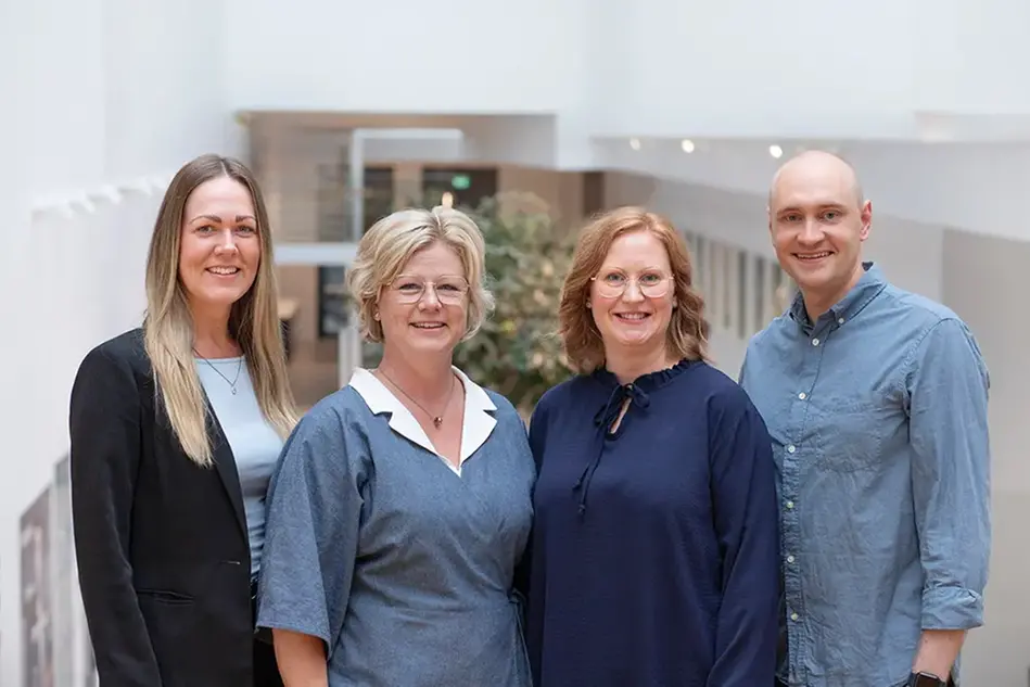 Tre kvinnor och en man står bredvid varandra. De ler och tittar in i kameran. Foto. 