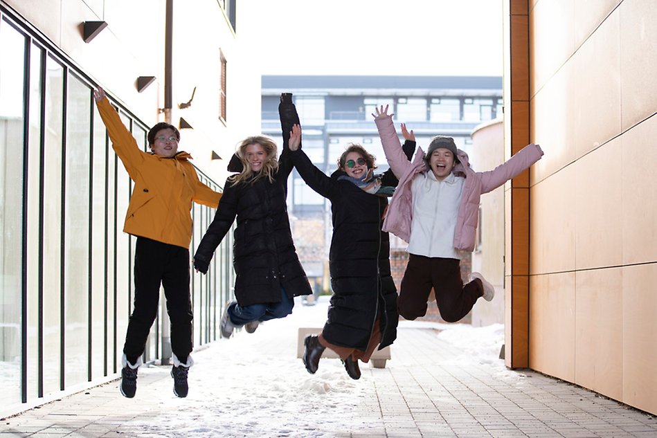 Fyra glada personer bredvid varandra hoppar i en utomhusmiljö.