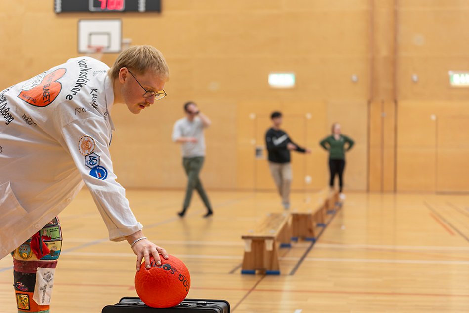 A person in a heavily decorated doctors coat reaching for a red ball. In the background a sports hall and a few people are visible. Photo.